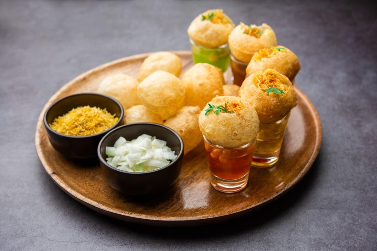 Crispy pani puri served with tangy flavored water in shot glasses accompanied by bowls of chopped onions and sev on a wooden plate