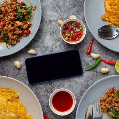 Which 25 Singapore Food Dishes Should You Try? 31 Top down view of Thai dishes including Pad Kra Pao with rice and Thai style omelets surrounding a blank smartphone