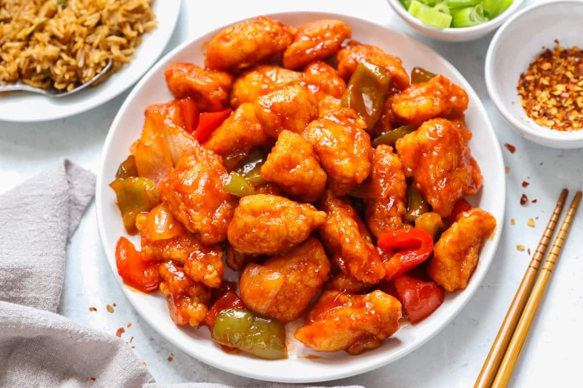 What Are 15 Chinese New Year Food Dishes You Must Try? 9 Sweet and sour chicken served on a white plate featuring crispy chicken pieces coated in glossy sauce with bell peppers and onions placed alongside chopsticks and side dishes