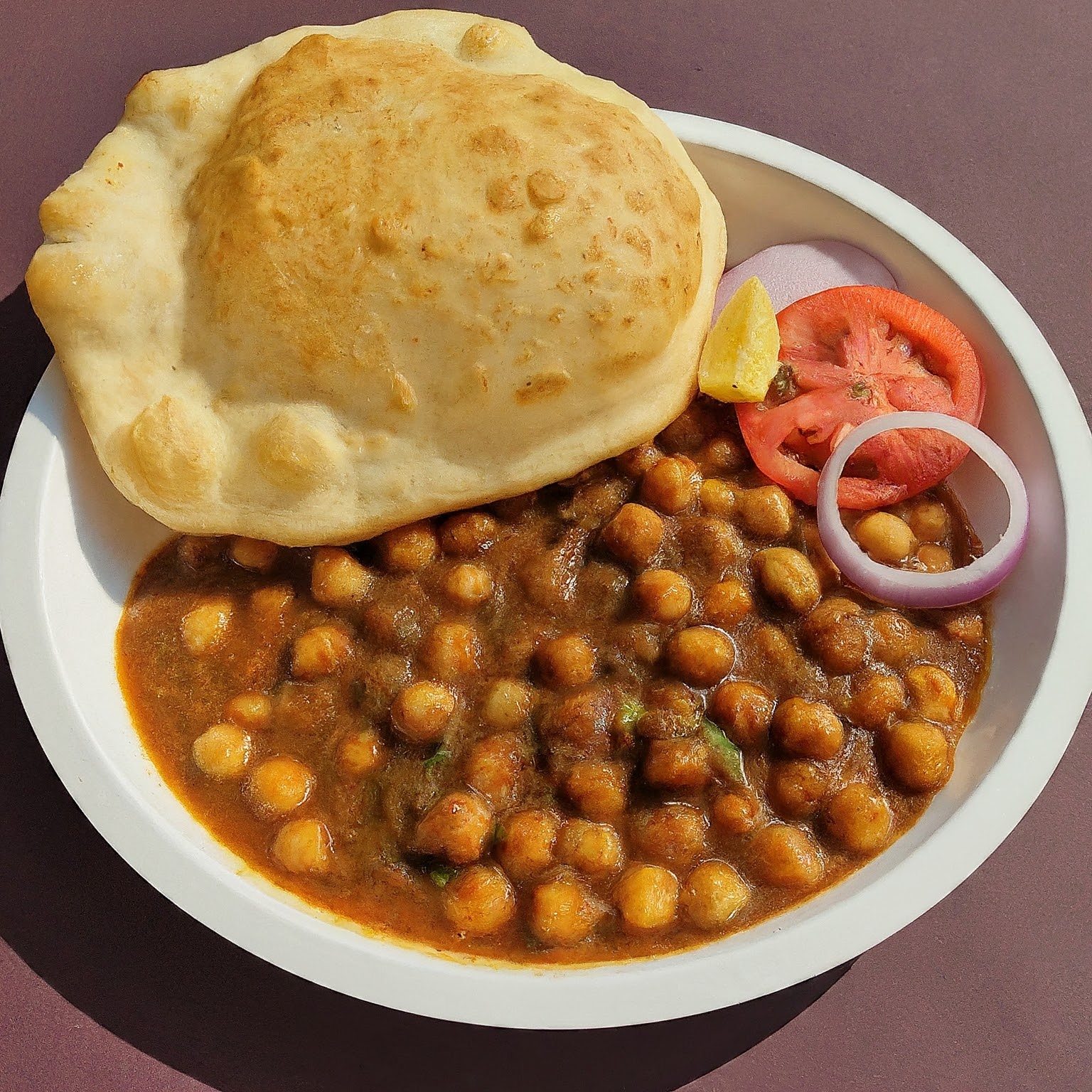 A plate of Chole Bhature with a bowl of red chickpea curry and a side of fluffy golden bhatura bread.