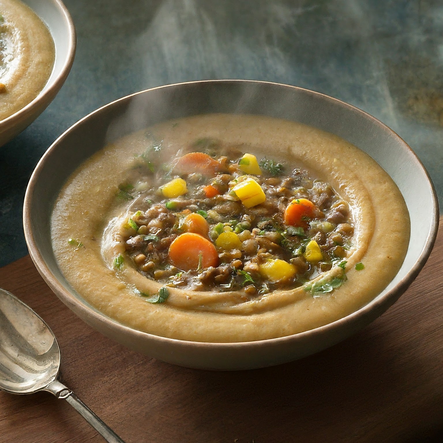 A steaming bowl of Hearty Millet and Lentil Soup with vegetables, herbs, and olive oil.