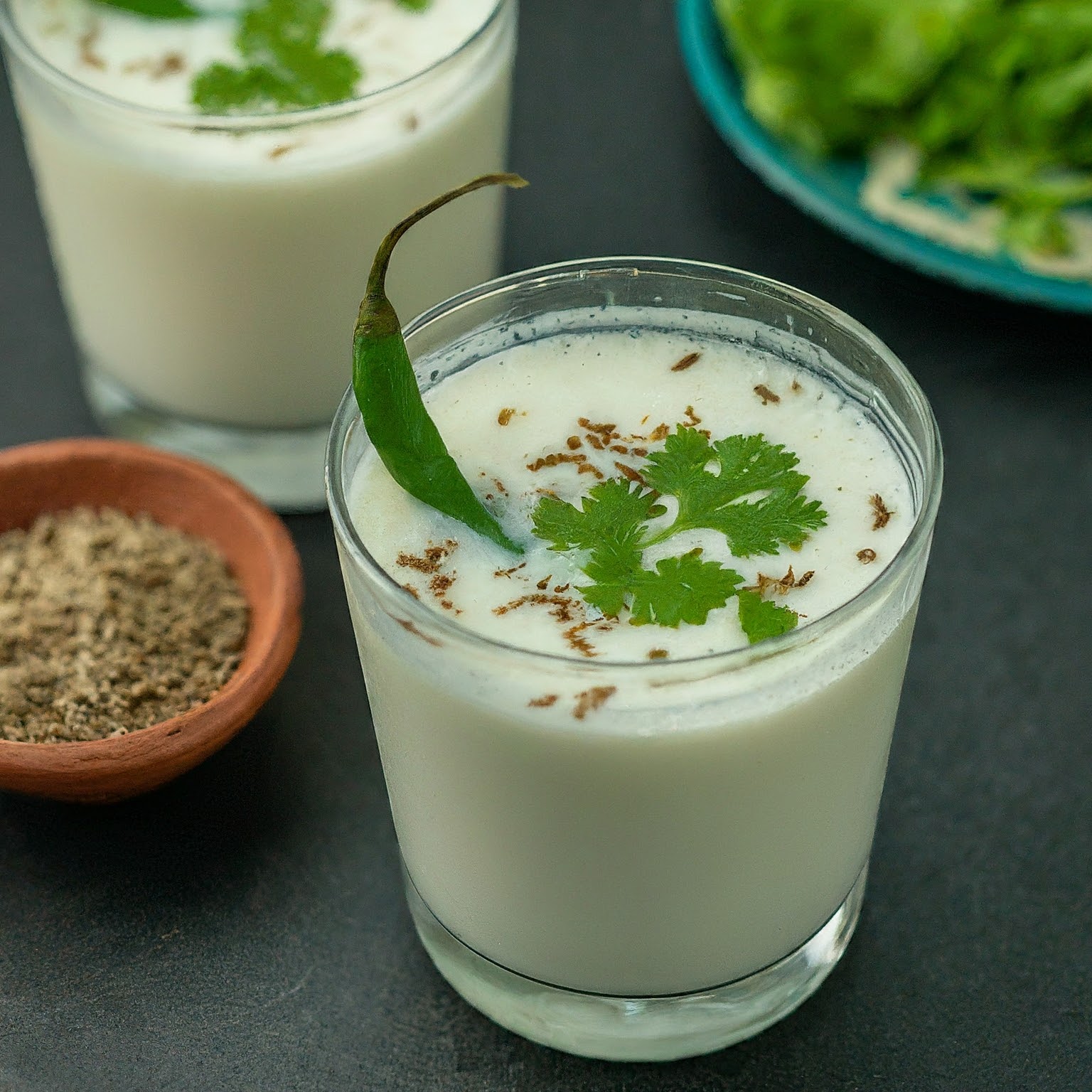 A tall glass of frothy buttermilk drink garnished with coriander leaves and a green chilli slice.