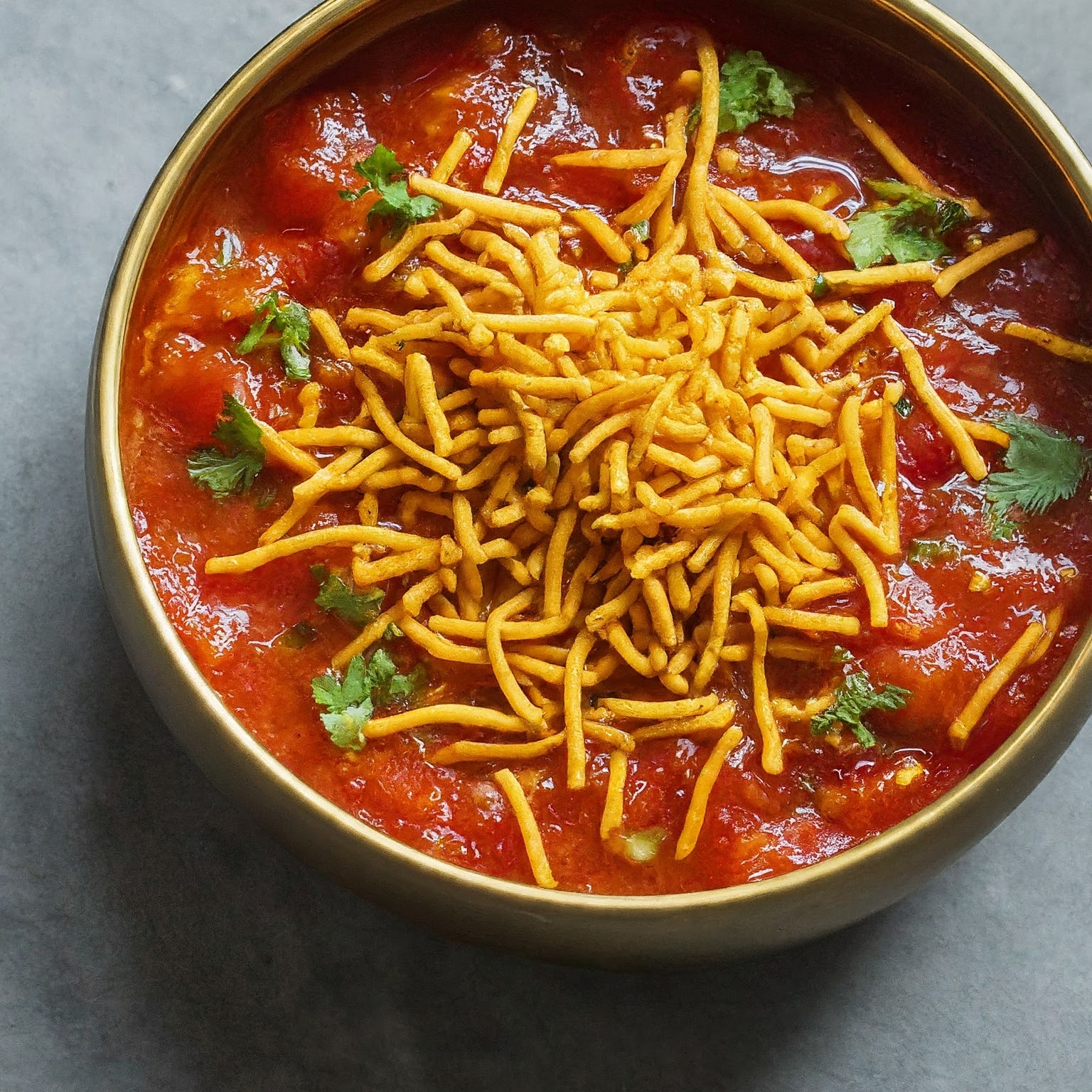 A round bowl filled with Gujarati Sev Tameta Nu Shaak, a tomato and sev noodle dish garnished with cilantro.